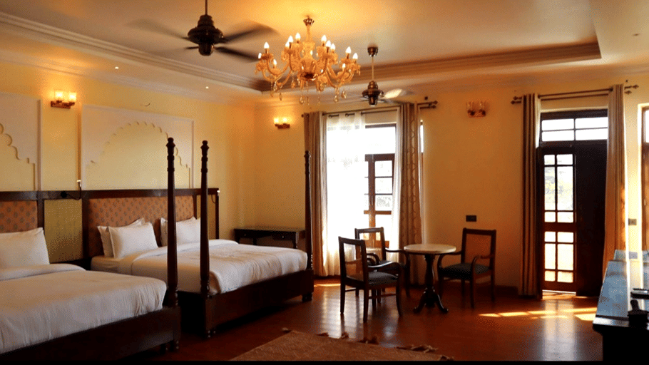 A room with a twin large bed, spreads, pillows, a fan, chairs, and a chandelier. | Hotel Devnadi, Haridwar