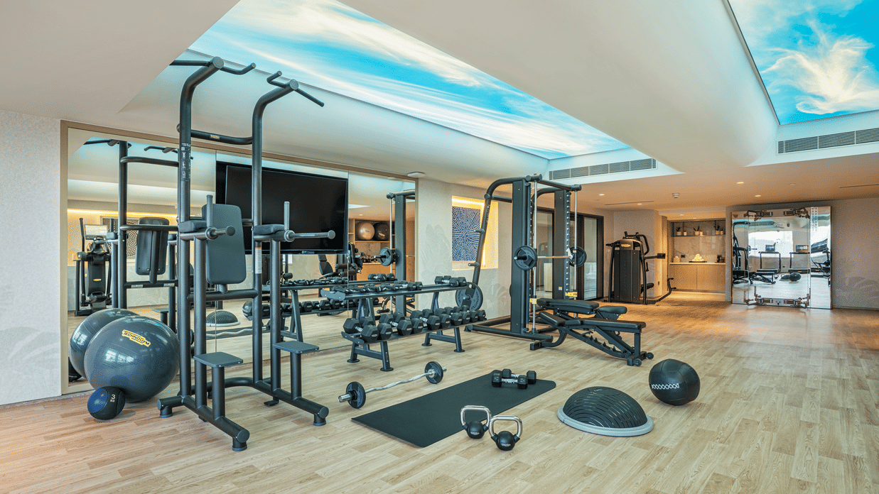 A gym at The Residency Towers Chennai featuring weight racks and exercise equipment with mirrored walls, and ceiling with the sky visible through the glass