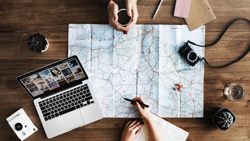 An image showing two hands going through a map on the table beside a laptop and a camera