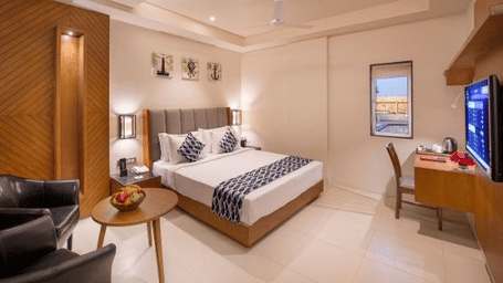 A cosy hotel room featuring a double bed with patterned cushions and a throw, a round side table with a fruit bowl, a black armchair, a wooden desk with a chair and a television at Lords Inn Motikhavdi.