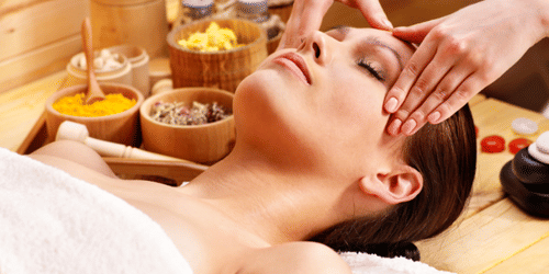 A woman lying with her eyes closed next to bowls of powdery substances, receiving massage on her head and temple at YO1 Longevity & Health Resorts