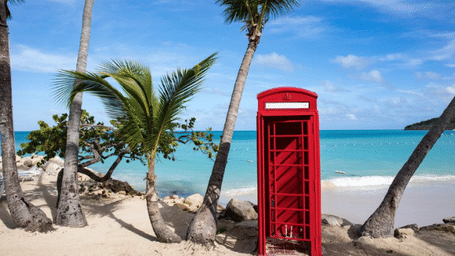 A vibrant red British telephone box stands on a white sand beach lined with palm trees, overlooking the turquoise ocean under a clear blue sky.