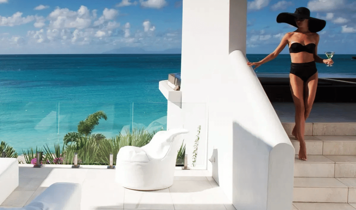 A woman in a black bikini and sun hat descends white stairs at a beachfront villa, holding a drink, with the blue sea beyond.