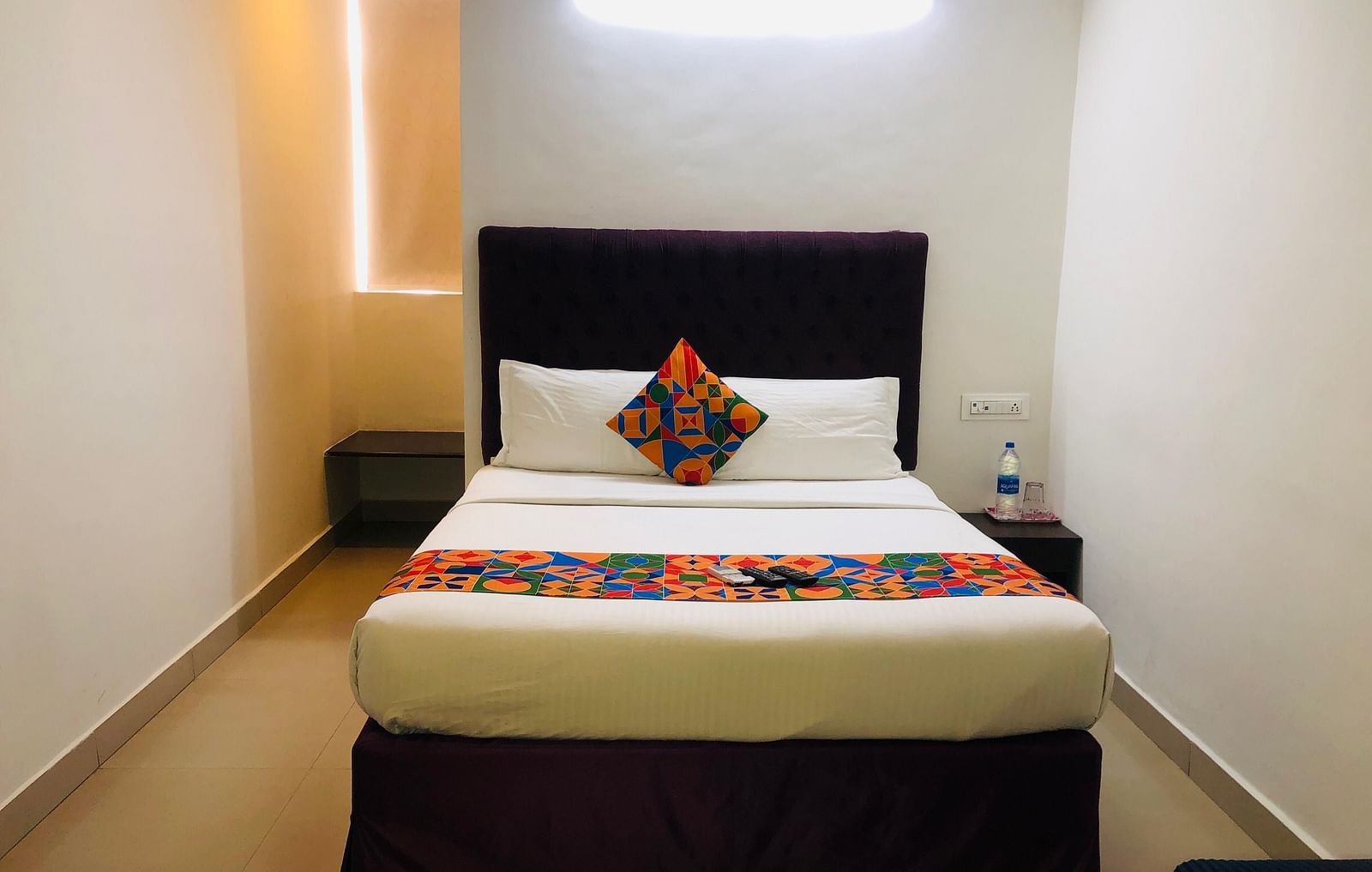 A neatly arranged hotel room with a double bed, a dark brown headboard, white linens, a decorative cushion, and a bedside table with a lamp - Astra Hotels and Suites, HSR Sector 7.