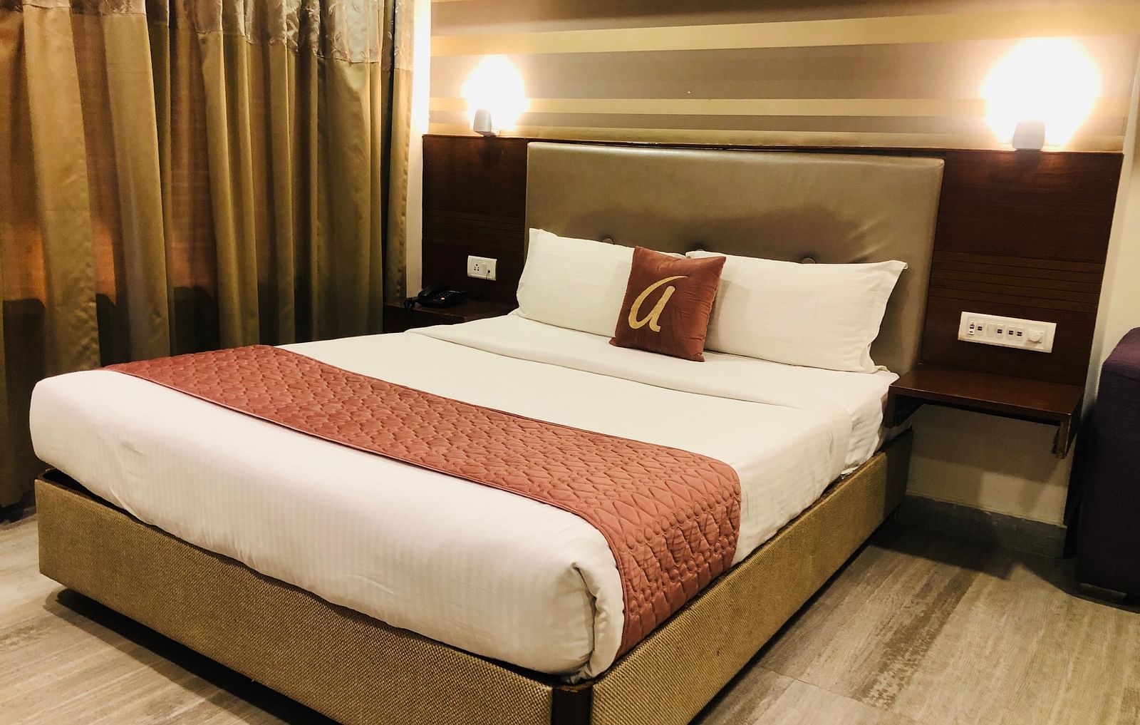 Hotel room double bed with white linens, maroon runner, and decorative pillows, illuminated by soft headboard lighting | Astra Hotels and Suites Kadubeesanahalli