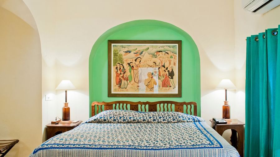 Bedroom with a bed featuring blue and white patterned bedding, bedside tables with lamps, artwork in a green alcove, teal curtains, and AC at Tijara Fort-Palace, Alwar.