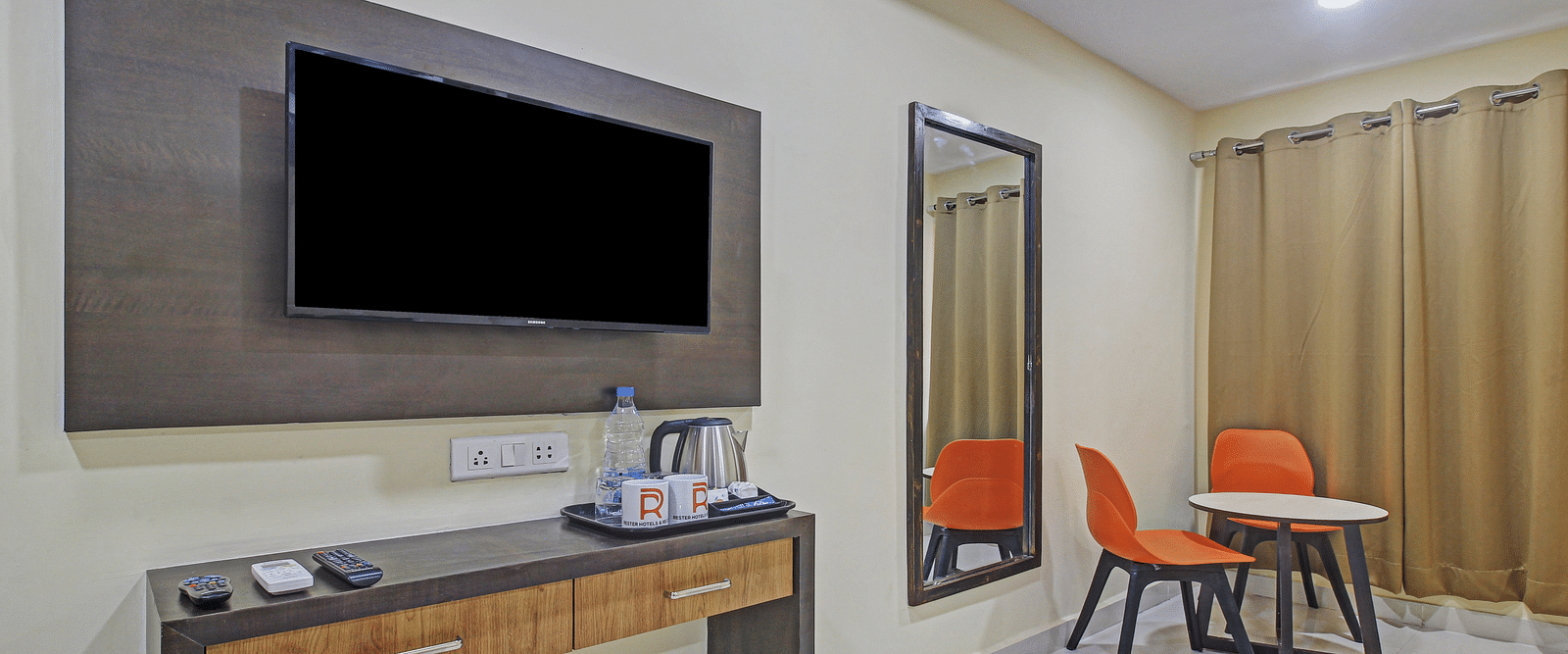 A neat hotel room with a wall-mounted TV, a wooden entertainment console, and a casual seating area with a vibrant orange chair.