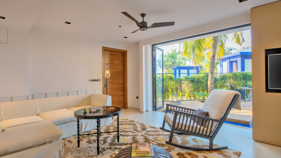 A living area of the room at Azaya Beach Resort Goa with a rocking chair, pouffe, table and couch facing the large door opening to the pool deck