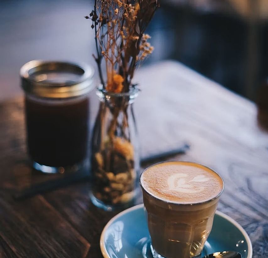 A cup of a coffee served on a blue coaster with a small vase holding dried flowers.