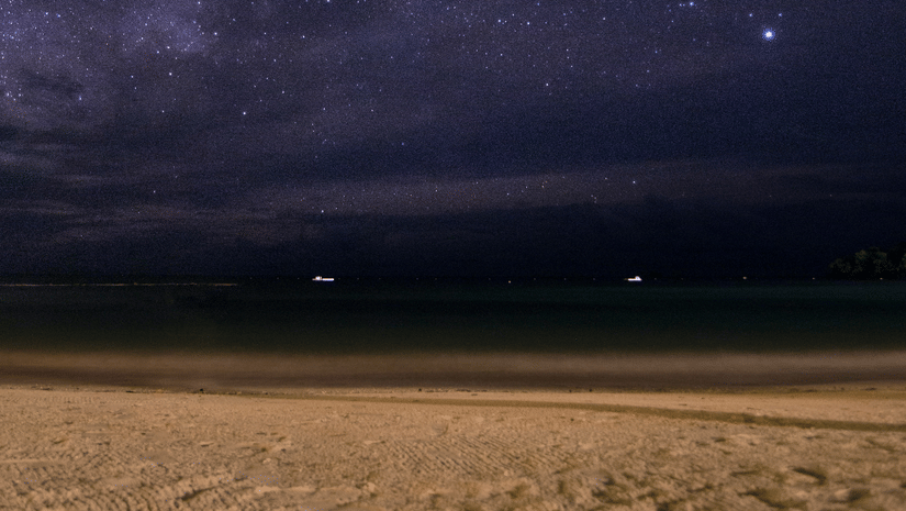 Star-filled night sky over a quiet beach, with the Milky Way stretching above calm seas and softly lit sandy shoreline