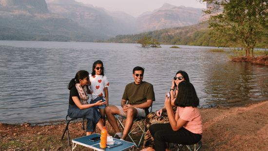 Four friends sitting in chairs by a lake enjoying a picnic, with hills and mountains in the hazy background.