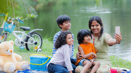 Mother taking a selfie with her three children while picnicking by a pond; a teddy bear and bike are nearby.