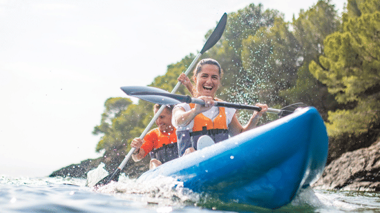 2 people smiling and paddling an inflatable blue kayak on a lake on a sunny day.