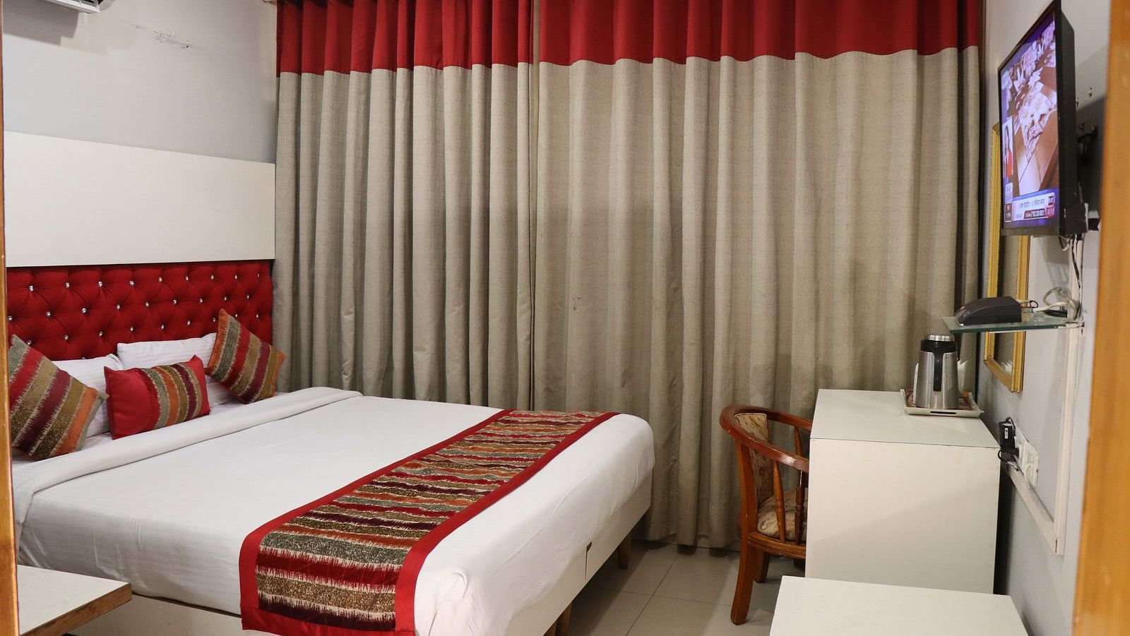 A hotel room with a plush bed, layered curtains with a red valance, a TV, a wooden chair, and an AC unit, illuminated by soft lighting. - Hotel Diamond Plaza, Chandigarh.