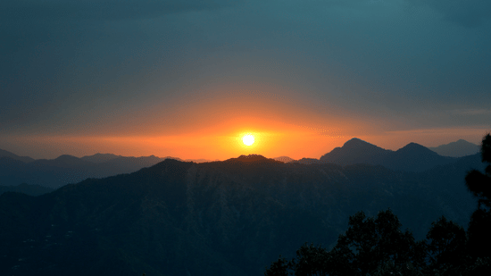 Sunset over the mountains of Kasauli, with the sun glowing above layered mountain peaks and silhouetted trees in the foreground.