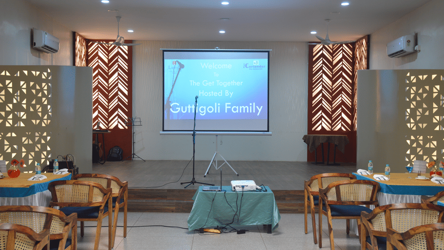 A banquet hall featuring a stage with a projector screen, and tables with a blue cloth - Lords Eco Inn Belagavi