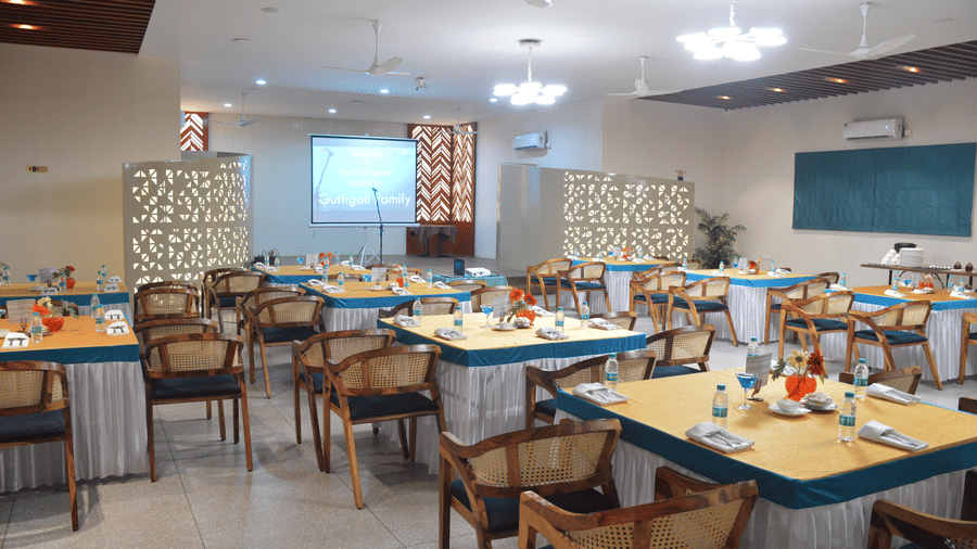 A banquet hall with a projector screen in the front with multiple tables and chairs under bright lights - Lords Eco Inn Belagavi