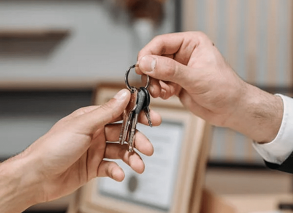 Two hands exchanging a set of keys over a table with a framed picture and other objects.