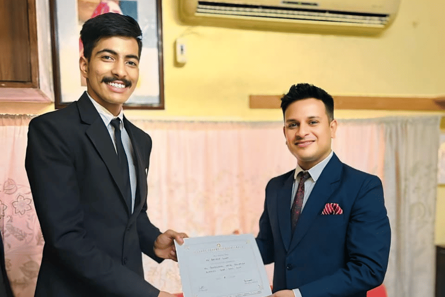 A person in a suit receives a certificate from another individual inside a room at Clarks Group of Hotels. They stand next to a sofa.