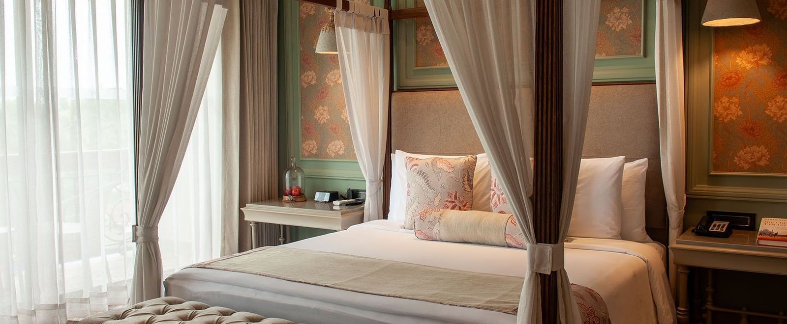  A luxurious suite room at The Evren, Vagator, with a four-poster bed draped in white curtains. The room features a decorative headboard wall with floral wallpaper and a tufted bench at the foot of the bed.