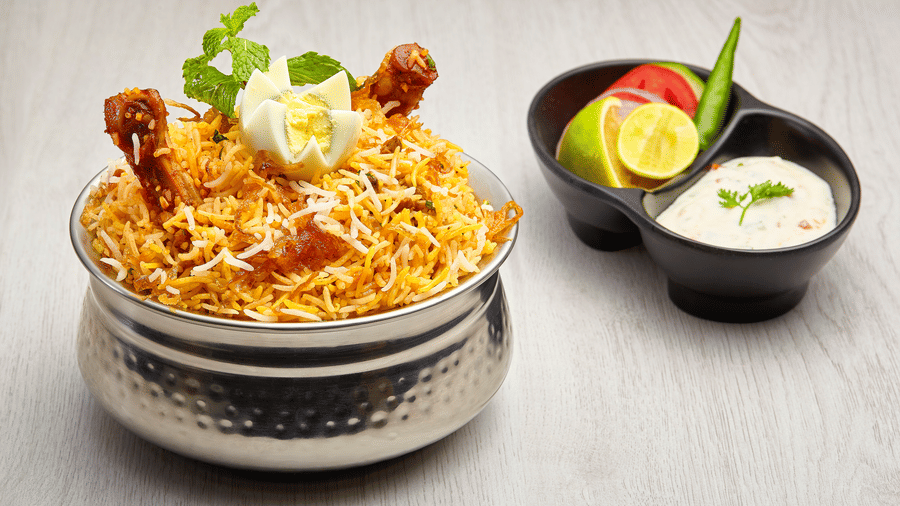 Hammered steel handi of chicken dum biryani garnished with mint and a boiled egg, served with a divided bowl of raita and salad.