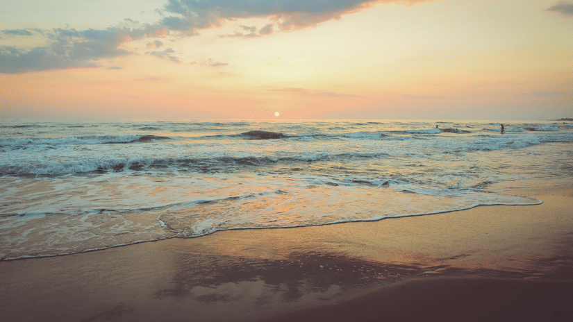 A view of ocean waves reaching the shore with the sky reflecting on the wet sand during sunset.