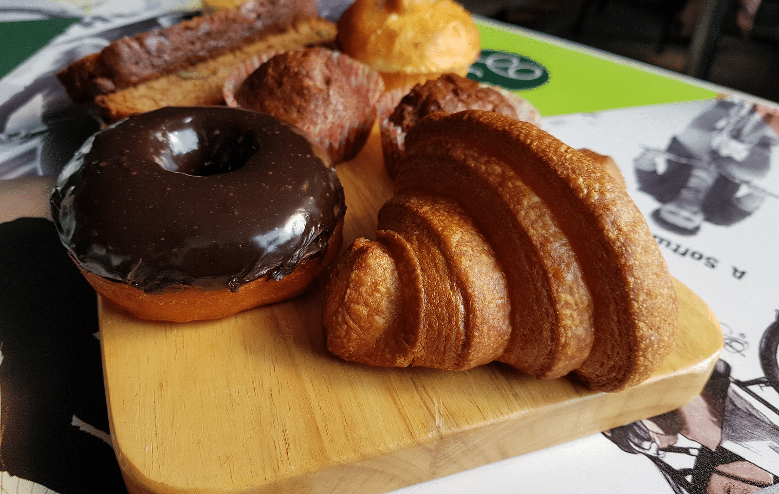 A variety of pastries on a wooden board, including a chocolate-glazed donut, a croissant, and several other baked goods
