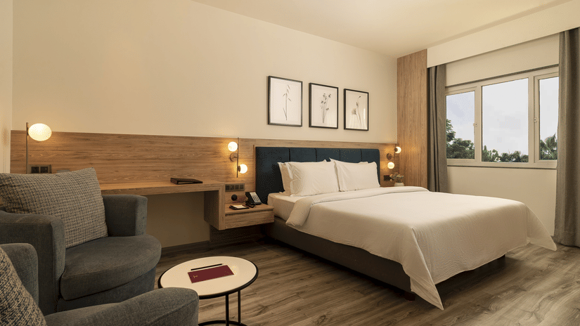 A warmly lit Premium Room features a double bed with white linens, two armchairs, a small round table, and framed pictures above the headboard at The Residency Karur.