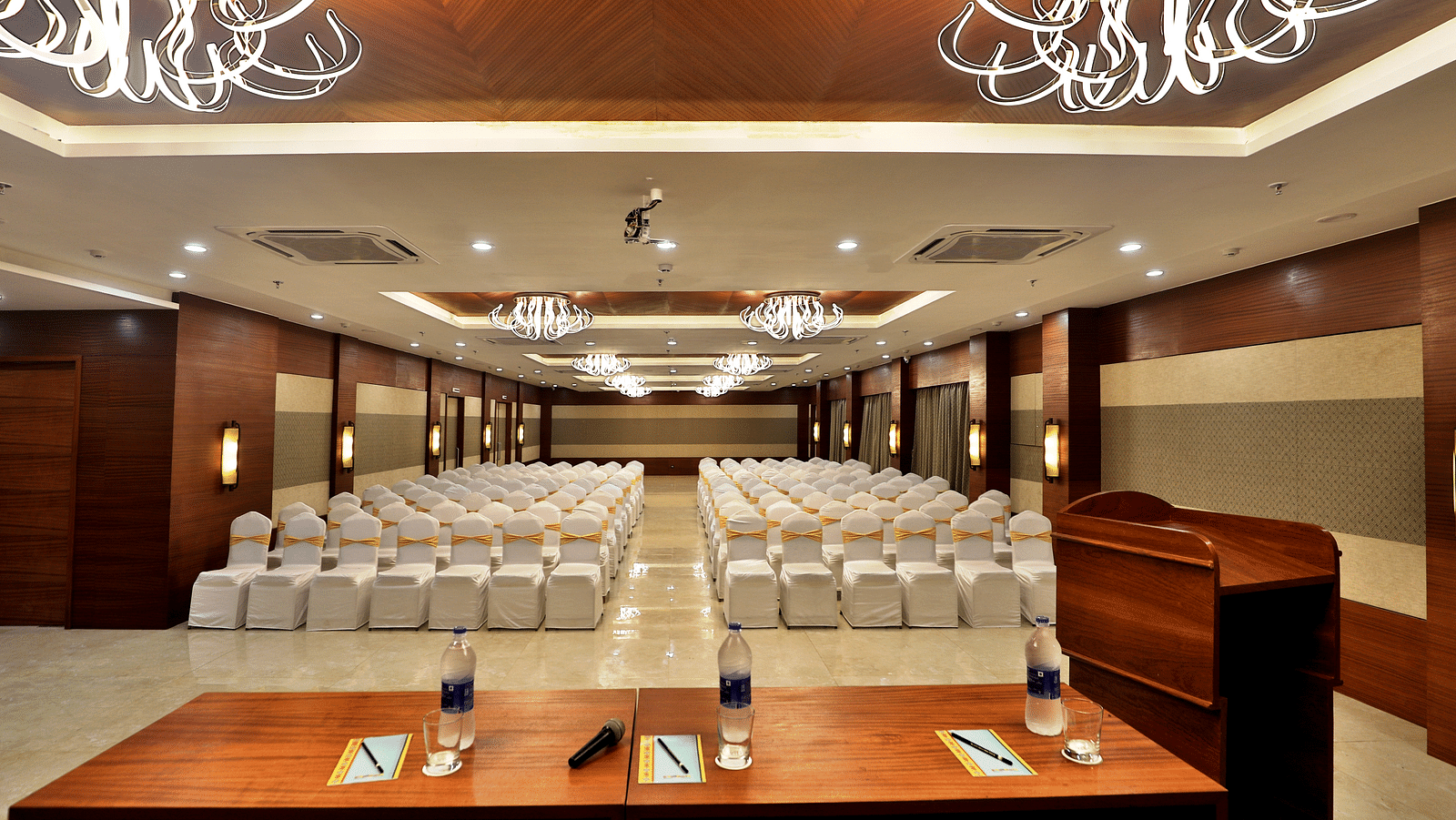 An image of a conference hall with numerous chairs and a table placed on the stage - Kamats India