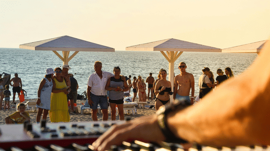 Close-up of hands playing an electronic keyboard with people and parasols on a beach in the background.