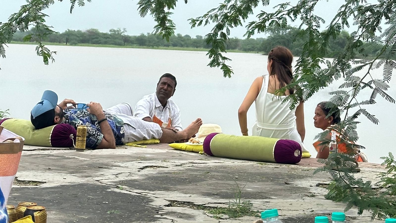 A group of individuals rests on mats by a water body, under the shade of leafy branches - provided by Fort Barli, Ajmer.