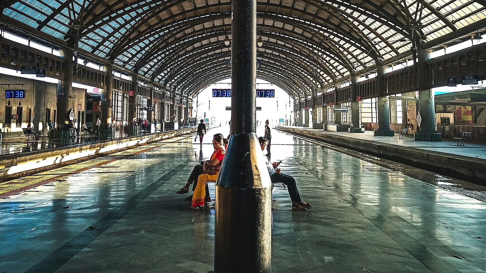 Two people sitting on a bench with their backs facing each other, one looking at the platform to the left, one looking at the right, at a railway station