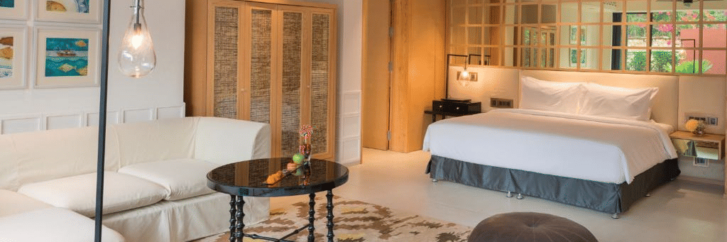 The private pool villa in Goa with a pouffe and a coffee table surrounded by a white couch in front of a plush bed at Azaya Beach Resort Goa