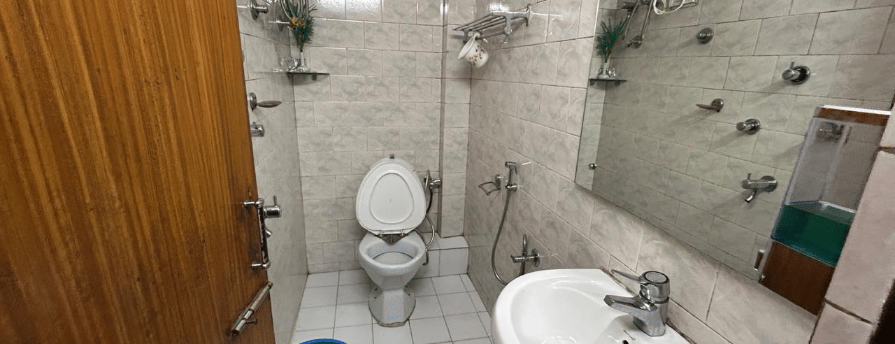 Bathroom with beige mosaic tiles, a large white sink, toilet, shower, and a blue bucket on the floor at Hotel Dolphin Darjeeling.