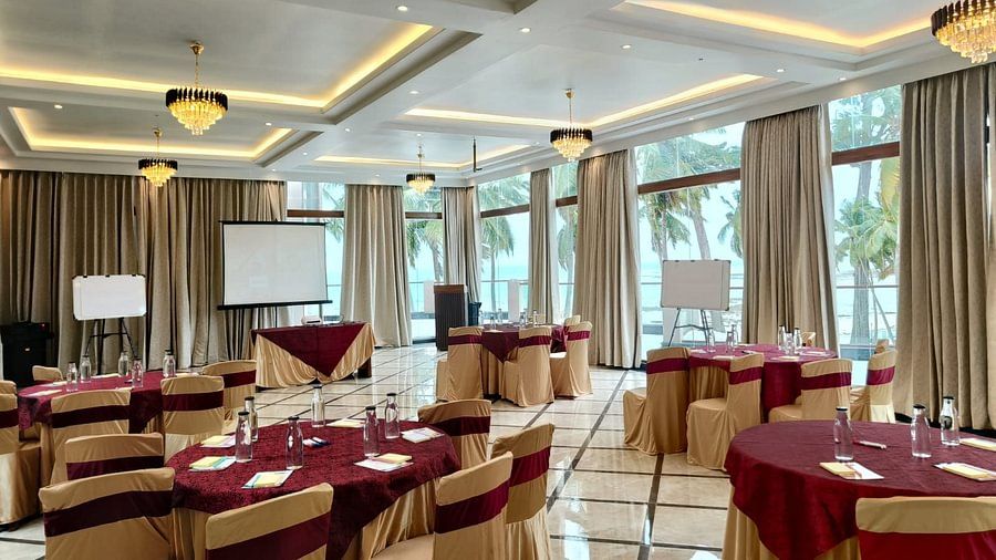 A large event space with neatly arranged round tables covered in beige and burgundy, illuminated by chandeliers, and large windows offering a view of palm trees outside.