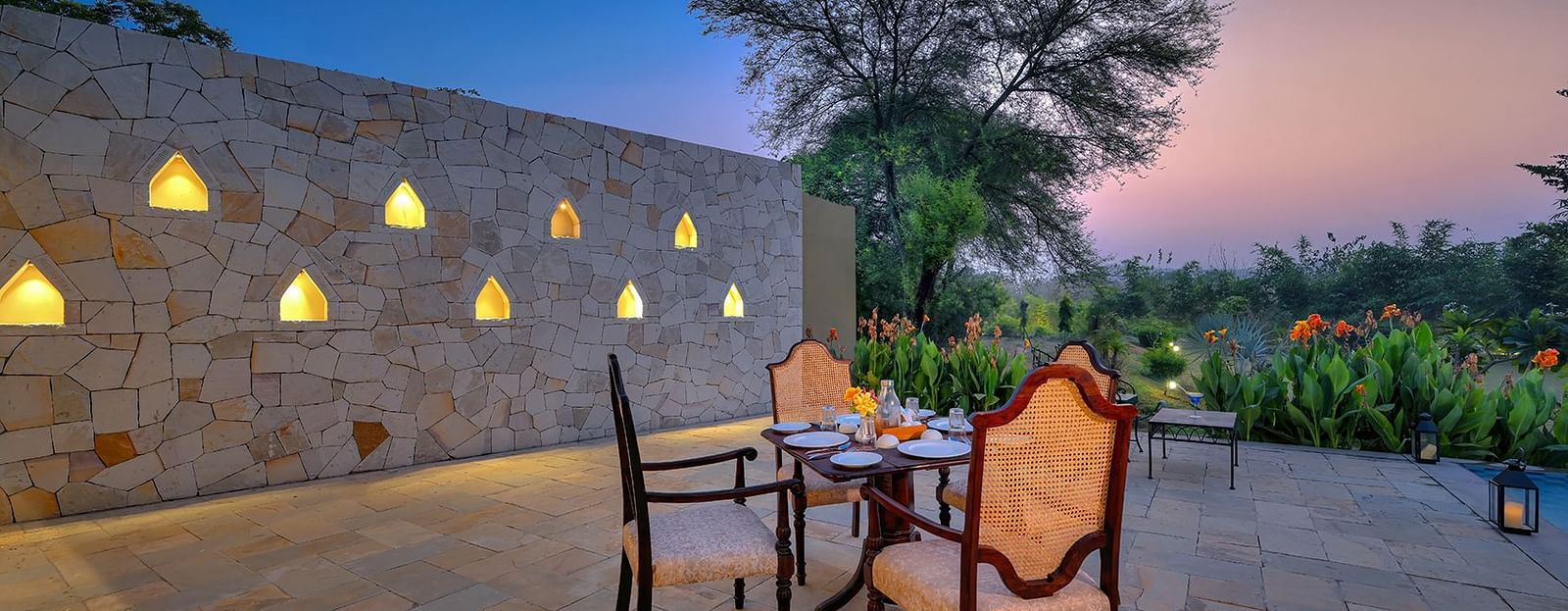 A dining table set with chairs, illuminated by decorative lights on the wall behind it - Singhvilas, Ranthambore