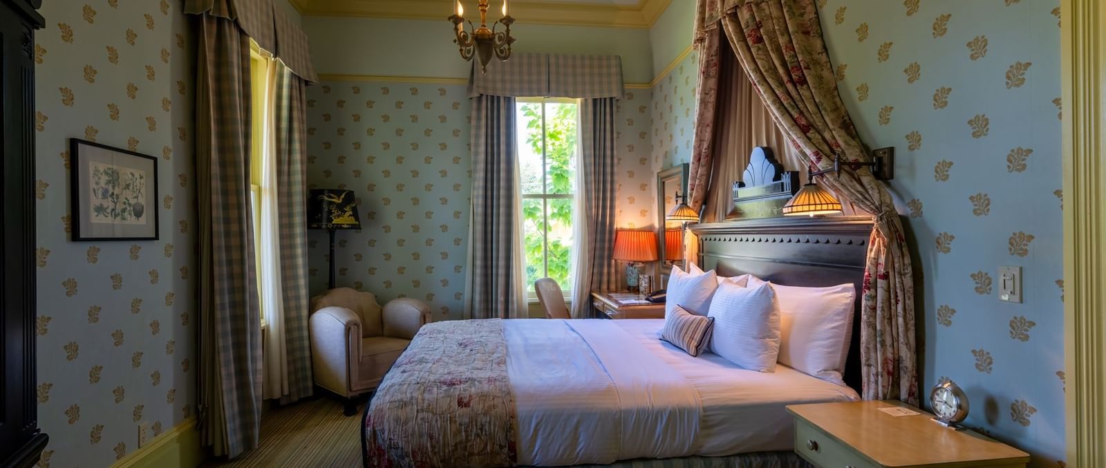 A spacious hotel bedroom with a comfortable bed, checkered bedspread, and a view to the outside at The Tallman Hotel.