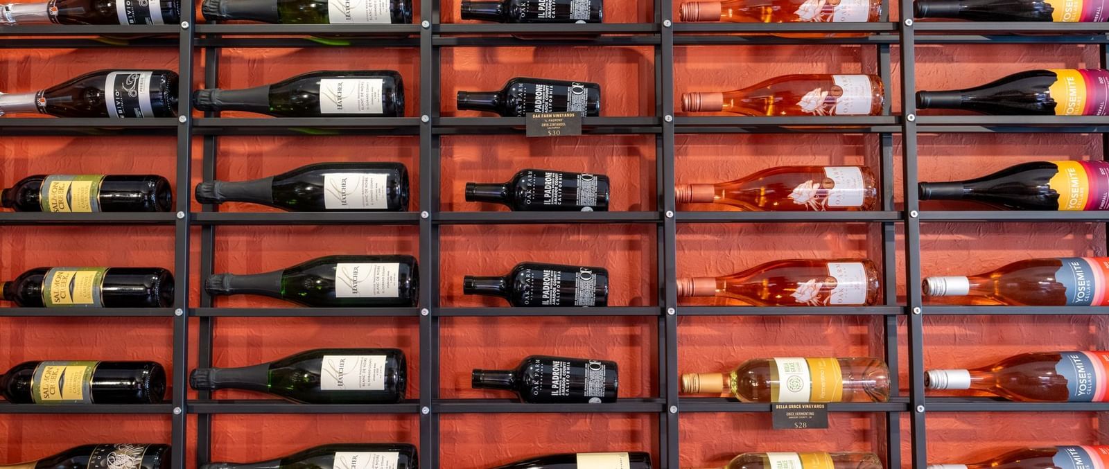  A wide shot of a large metal wine rack, displaying multiple rows of wine bottles on a red wall at Aroma Restaurant and Wine Bar at The Groveland Hotel.