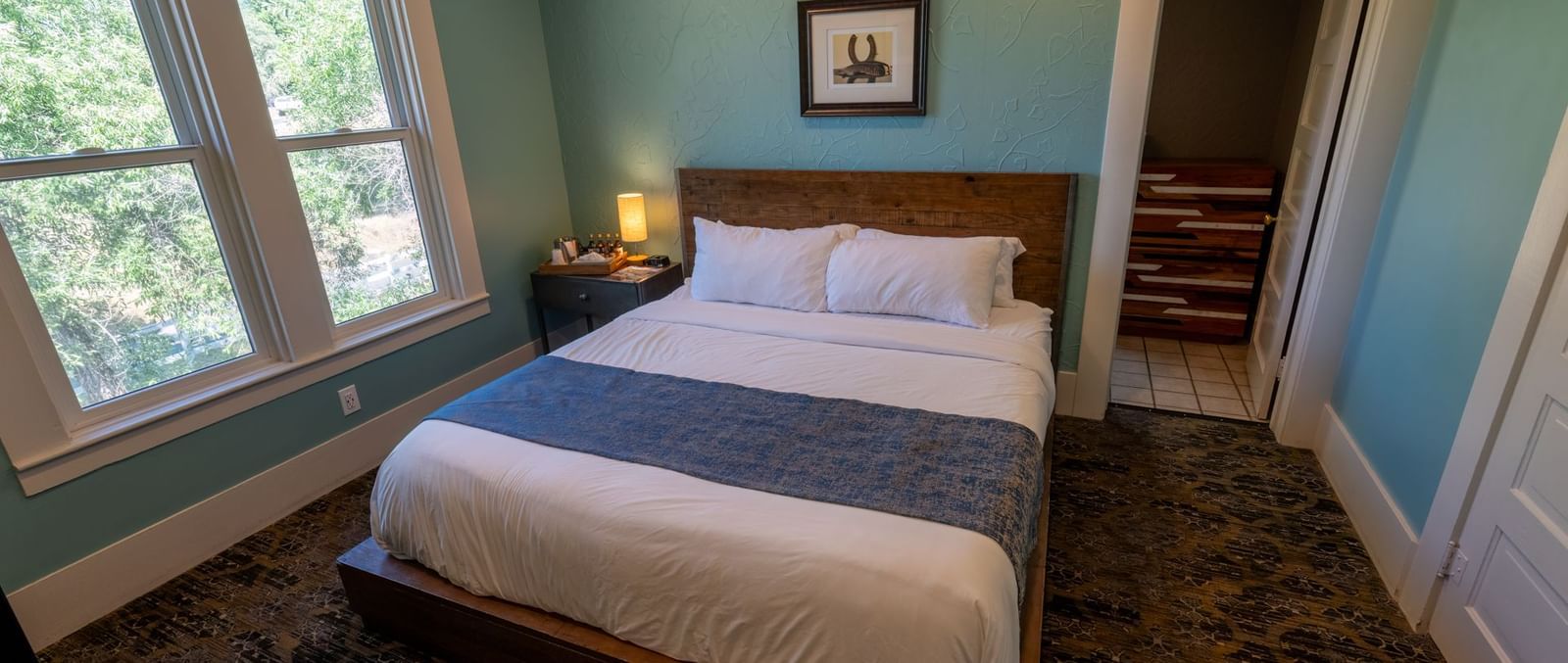 Corner view of a Family Suite at The Groveland Hotel featuring a plush bed, carpeted flooring, and a wood-framed glass window offering a view of the greenery nearby.