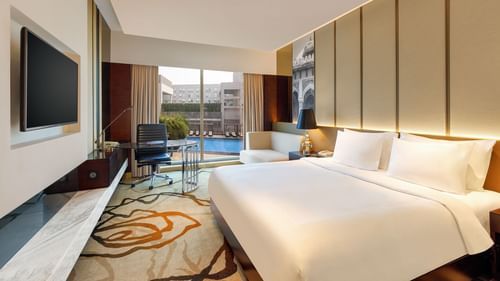 Premium King Room at Pride Plaza Hotel in New Delhi featuring a king bed in front of a TV mounted on the wall next to which a work desk is placed with a backdrop of pool view