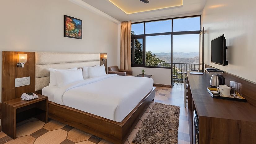 Jungle View Deluxe Room at Pride Ashiyana Resort Mussoorie featuring a plush bed facing the TV mounted on the wall and coffee maker with cups on the table below it. In the background a large window touching the floor and the roof is boasting a majestic vie
