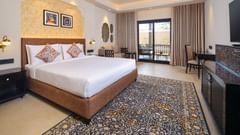  A luxurious hotel room at Pride Amber Vilas Resort Jaipur, featuring a large bed with ornate headboard, two nightstands with lamps, a patterned rug, a seating area, and a balcony with outdoor views.