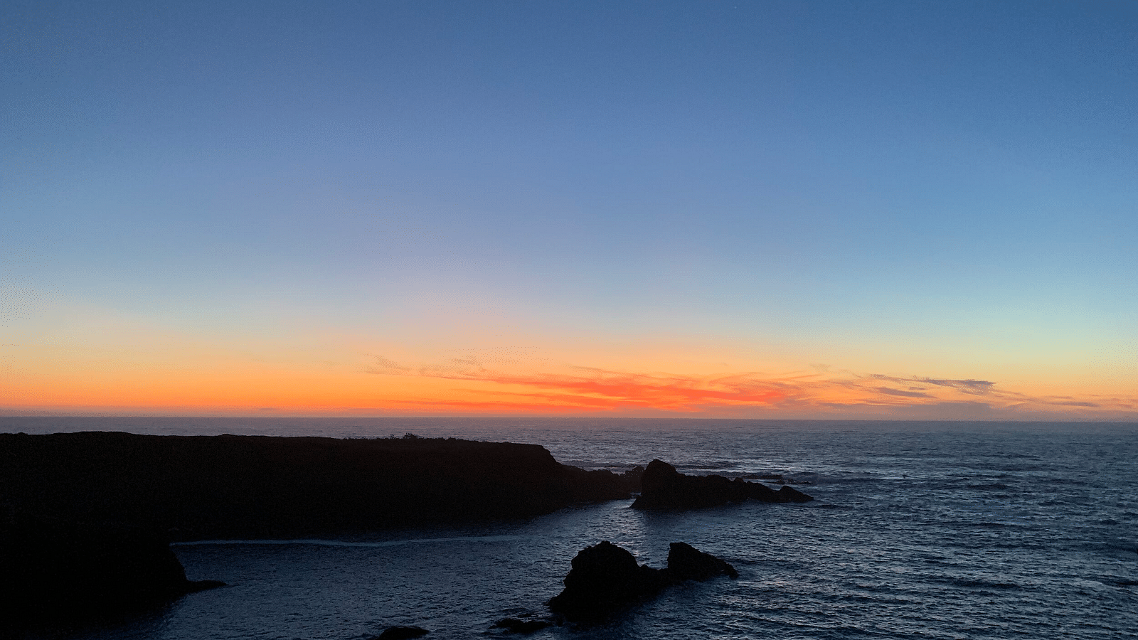 A twilight scene with a gradient sky above a dark silhouette of a rocky coastline meeting the ocean.