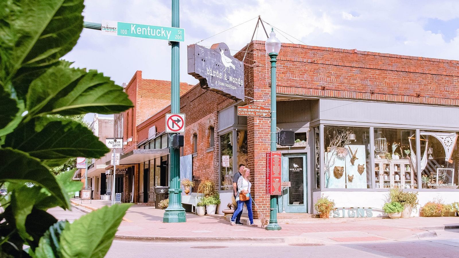 A crossing with a marketplace with brick like textures on the exterior with physical signs to show directions, a couple walking on the road and green leafs on the side.