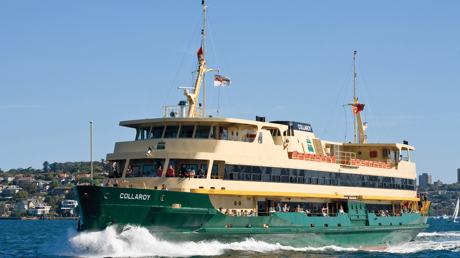 Green and cream passenger ferry named Collaroy cutting through blue harbor water past a tree-lined residential shoreline on a clear sunny day