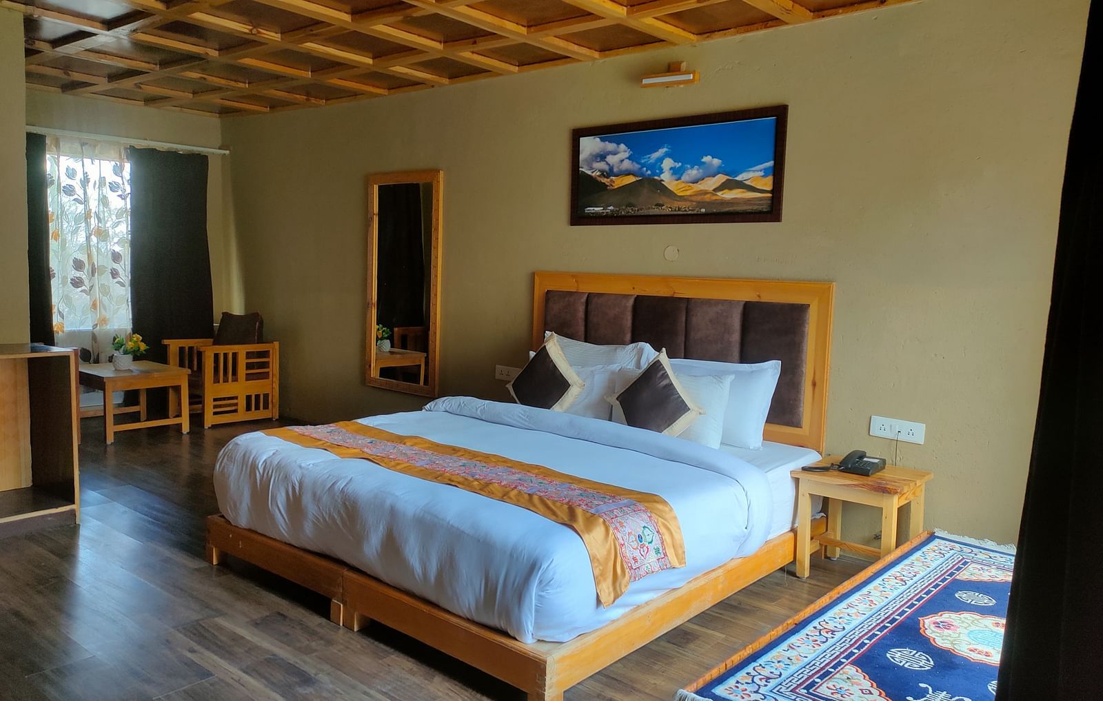 Deluxe Cottage showcases a neatly made king size bed with a striped throw, accompanied by wooden furniture, a decorative ceiling, and a framed picture at White Sands Resort, Ladakh.