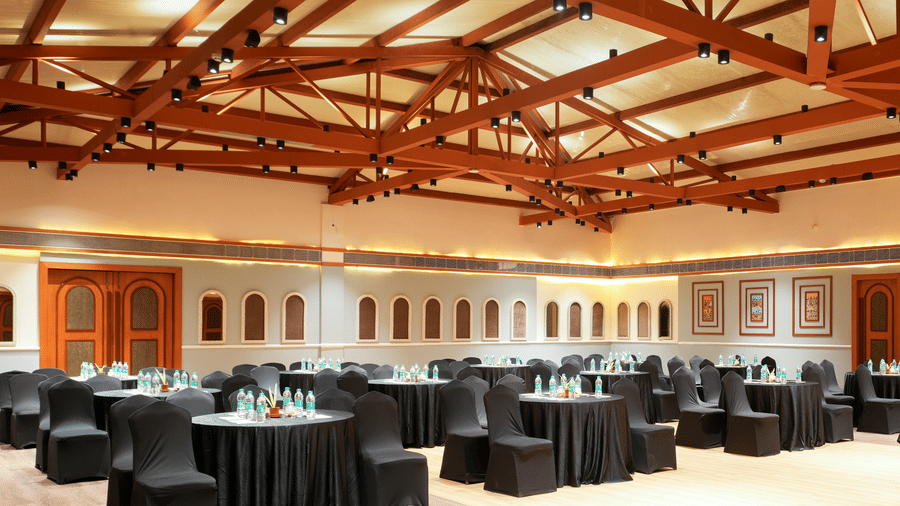 Banquet hall at Vedic Village Spa Resort Kolkata with exposed wooden truss ceiling, round tables, black chair covers, and arched windows along the walls.