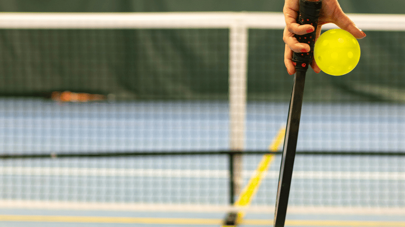 A hand holding a black pickleball paddle and a yellow ball by a court net