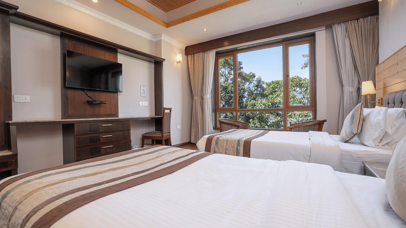 A hotel room with a double bed, a wall-mounted television, and a window draped with brown and sheer white curtains.