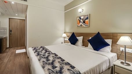 Premium Studio at Starlit Suites Newtown, Kolkata features a double bed with a blue and white patterned throw and cushions, bedside tables with lamps, and a doorway.